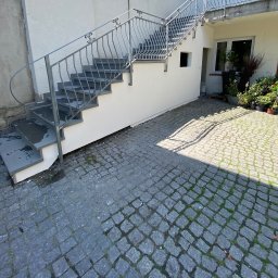 KUB - HOUSE Jakub Piechowicz - Zewnętrzne schody z metalową balustradą prowadzące na górę, świeżo pomalowane, z mokrymi stopniami i kostką brukową na dziedzińcu.