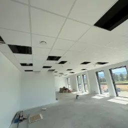 Nowoczesne wnętrze biurowe z podwieszanym sufitem kasetonowym, częściowo zamontowanym, z widocznymi otworami na instalacje. Duże okna wpuszczają naturalne światło.