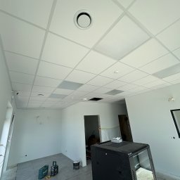 KUB - HOUSE Jakub Piechowicz - Biały sufit kasetonowy z okrągłym nawiewnikiem w nowoczesnym biurze, szare płytki podłogowe, narzędzia i szafa rack w tle, remont w trakcie.