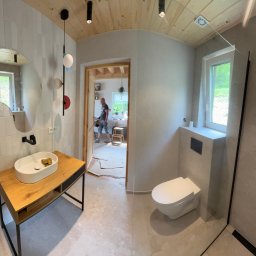 KUB - HOUSE Jakub Piechowicz - Wykończona łazienka z drewnianym sufitem, umywalką na drewnianym blacie i kabiną prysznicową. W tle widoczny fragment innego pomieszczenia w trakcie remontu.