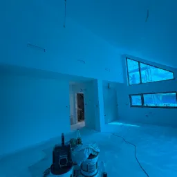 Wnętrze po malowaniu: białe ściany, okna z widokiem na pracownika na rusztowaniu. Na pierwszym planie wiaderka z farbą i narzędzia malarskie.