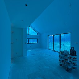 KUB - HOUSE Jakub Piechowicz - Wnętrze po gładziowaniu, przygotowane do malowania. Widoczne ściany, okna, puszki elektryczne i wiaderka z farbą. Skosy sufitu i duże okno dachowe doświetlają pomieszczenie.