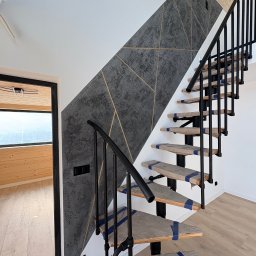 KUB - HOUSE Jakub Piechowicz - Ściana z dekoracyjną szarą strukturą i złotymi liniami, schody z czarną balustradą zabezpieczone taśmą malarską, widok na drewniane wnętrze przez otwarte drzwi.