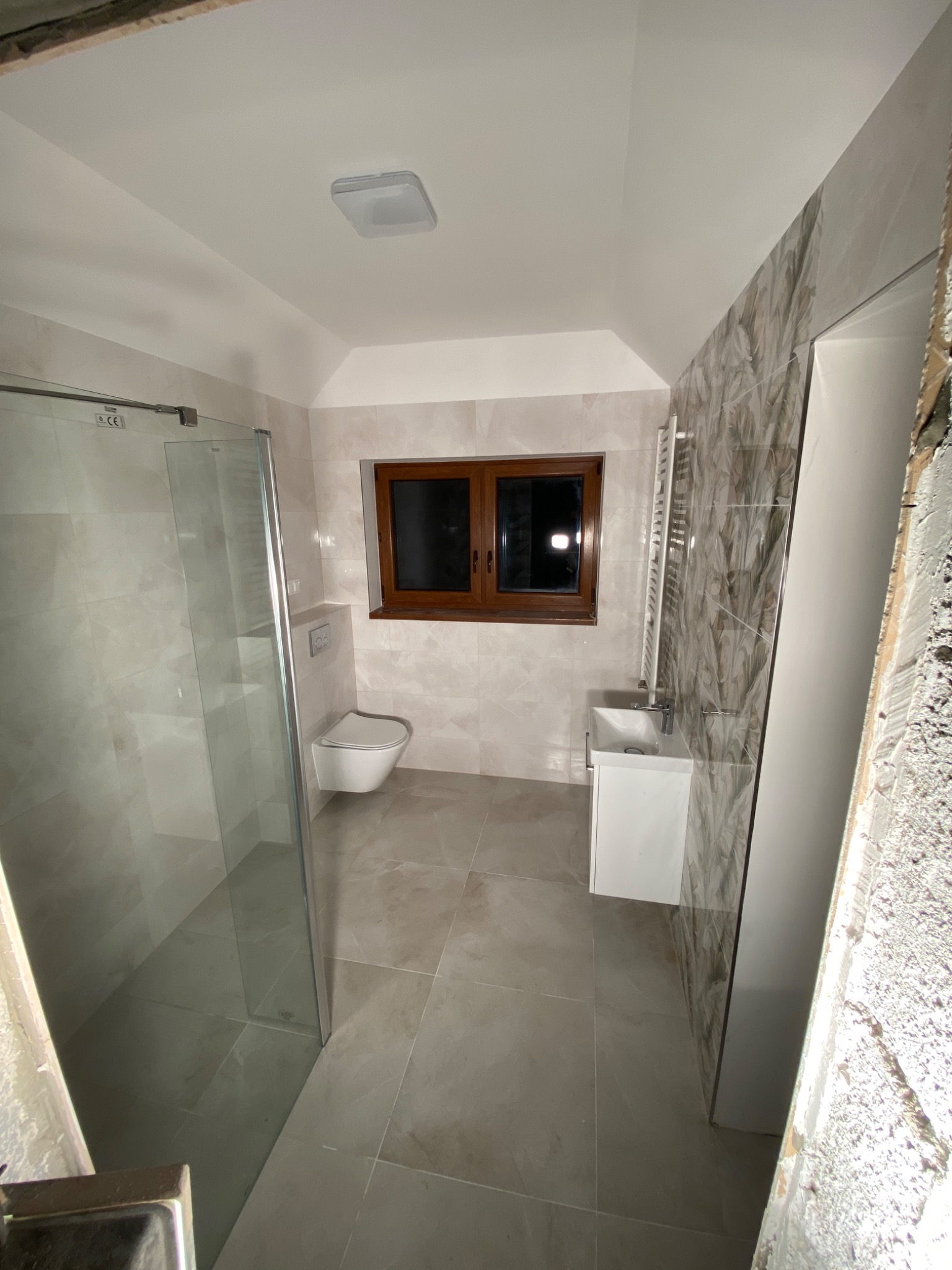 Wykończona łazienka z kabiną prysznicową, podwieszaną toaletą i umywalką, płytki imitujące kamień na ścianach i podłodze, okno z brązową ramą, widoczne prace wykończeniowe przy drzwiach.