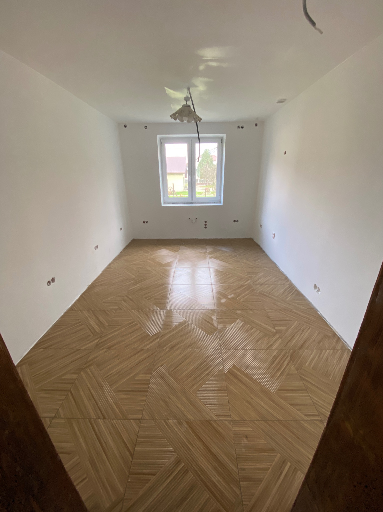 Pusty pokój po remoncie z ułożoną parkietową podłogą z płytek, białe ściany z widocznymi gniazdkami, okno z widokiem na ogród i wisząca lampa bez klosza.