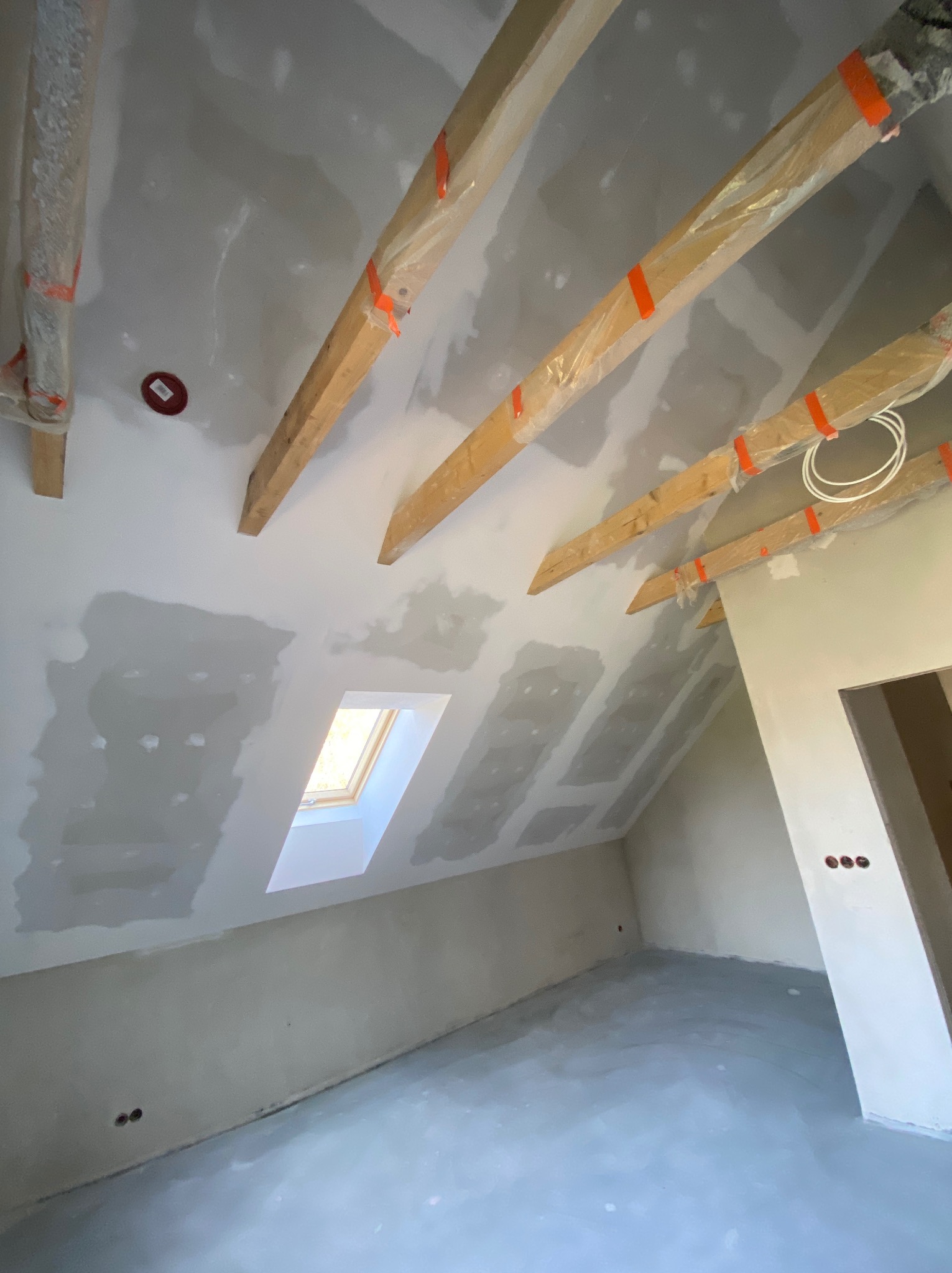 Poddasze w trakcie prac wykończeniowych: widoczne drewniane belki zabezpieczone folią, okno dachowe, ściany pokryte szpachlą w trakcie nakładania warstwy, przygotowane pod gładzie, oraz surowa...
