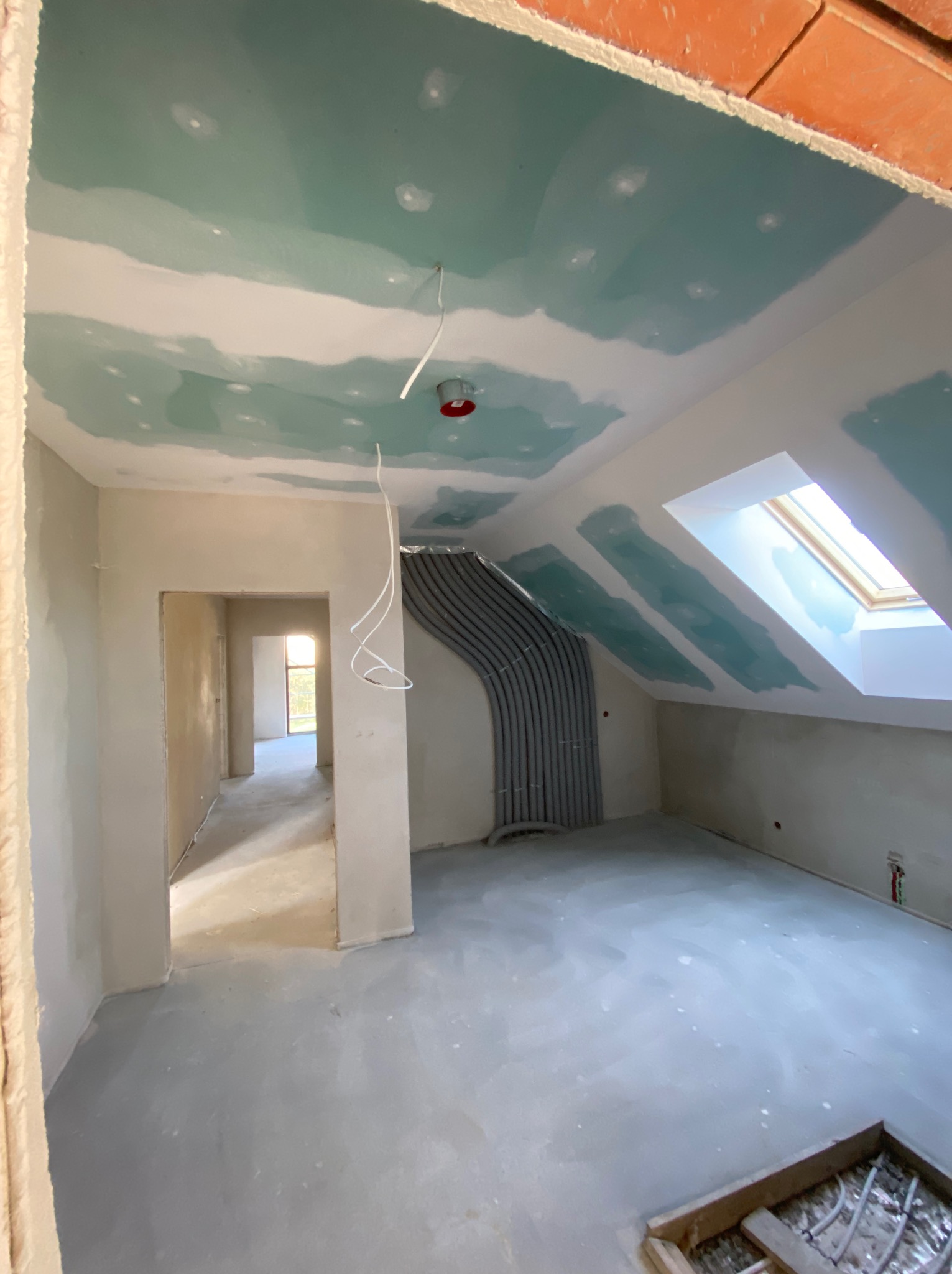 Poddasze w trakcie prac wykończeniowych: ściany i sufit pokryte wstępną warstwą gładzi, widoczne rury instalacji, okno dachowe i otwór drzwiowy do kolejnego pomieszczenia.