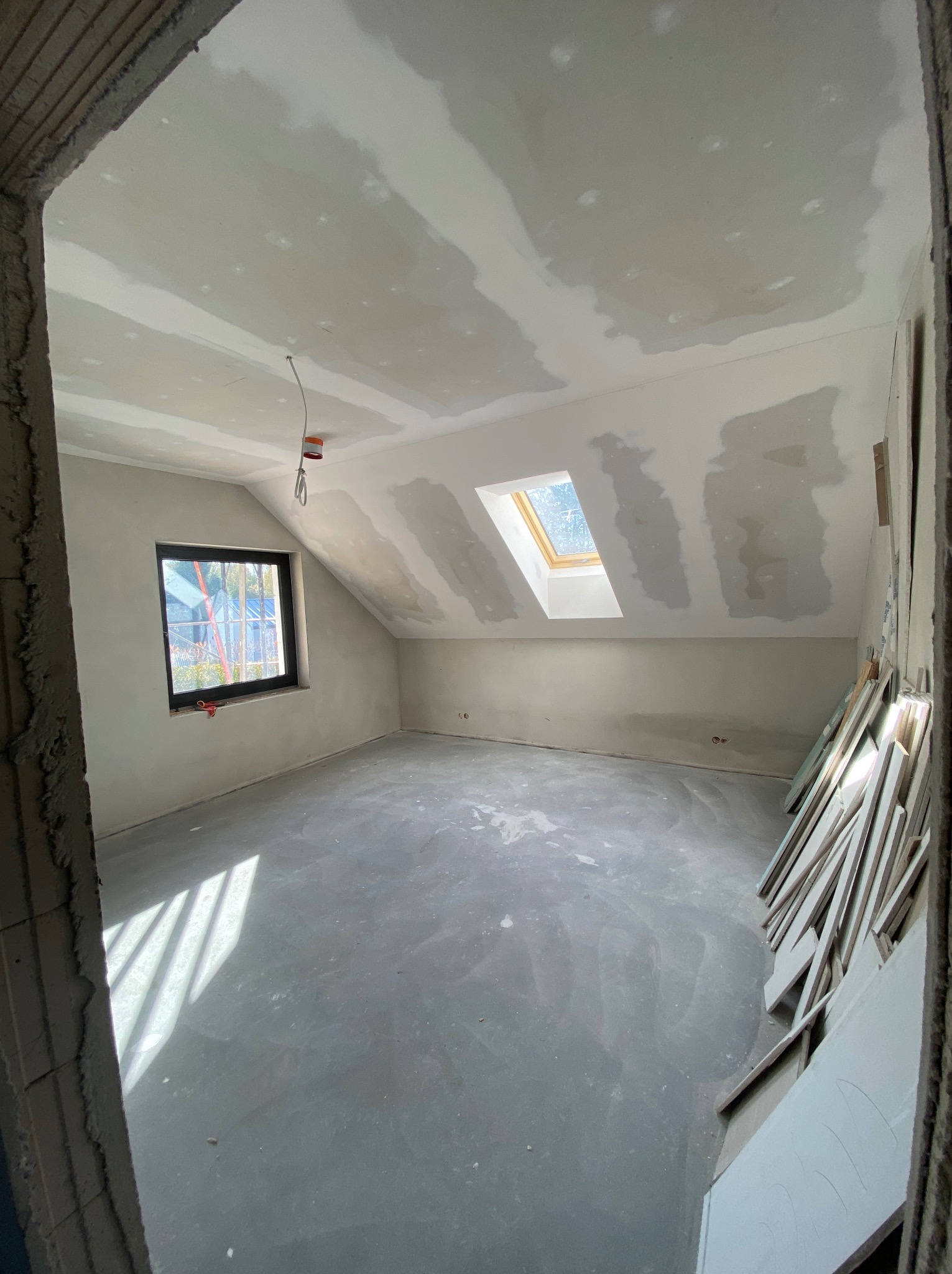 Poddasze w trakcie prac wykończeniowych: ściany i sufit pokryte wstępną warstwą gładzi, widoczne łączenia płyt kartonowo-gipsowych, okno dachowe i okno w ścianie szczytowej, na podłodze stos płyt K-G.