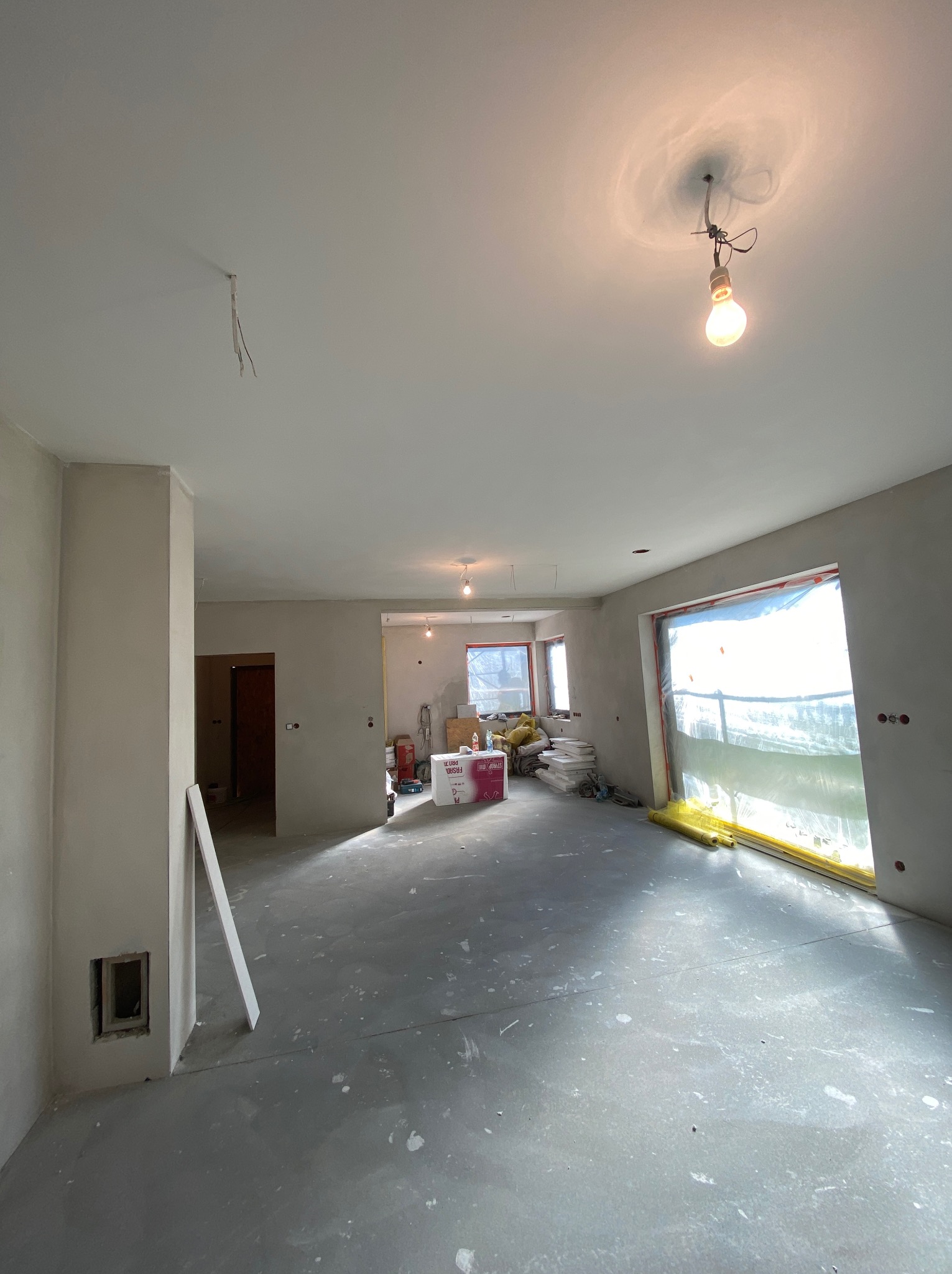 Wnętrze w trakcie prac wykończeniowych: ściany pokryte świeżo nałożoną warstwą gładzi, okna zabezpieczone folią malarską, widoczne materiały budowlane i surowa żarówka.
