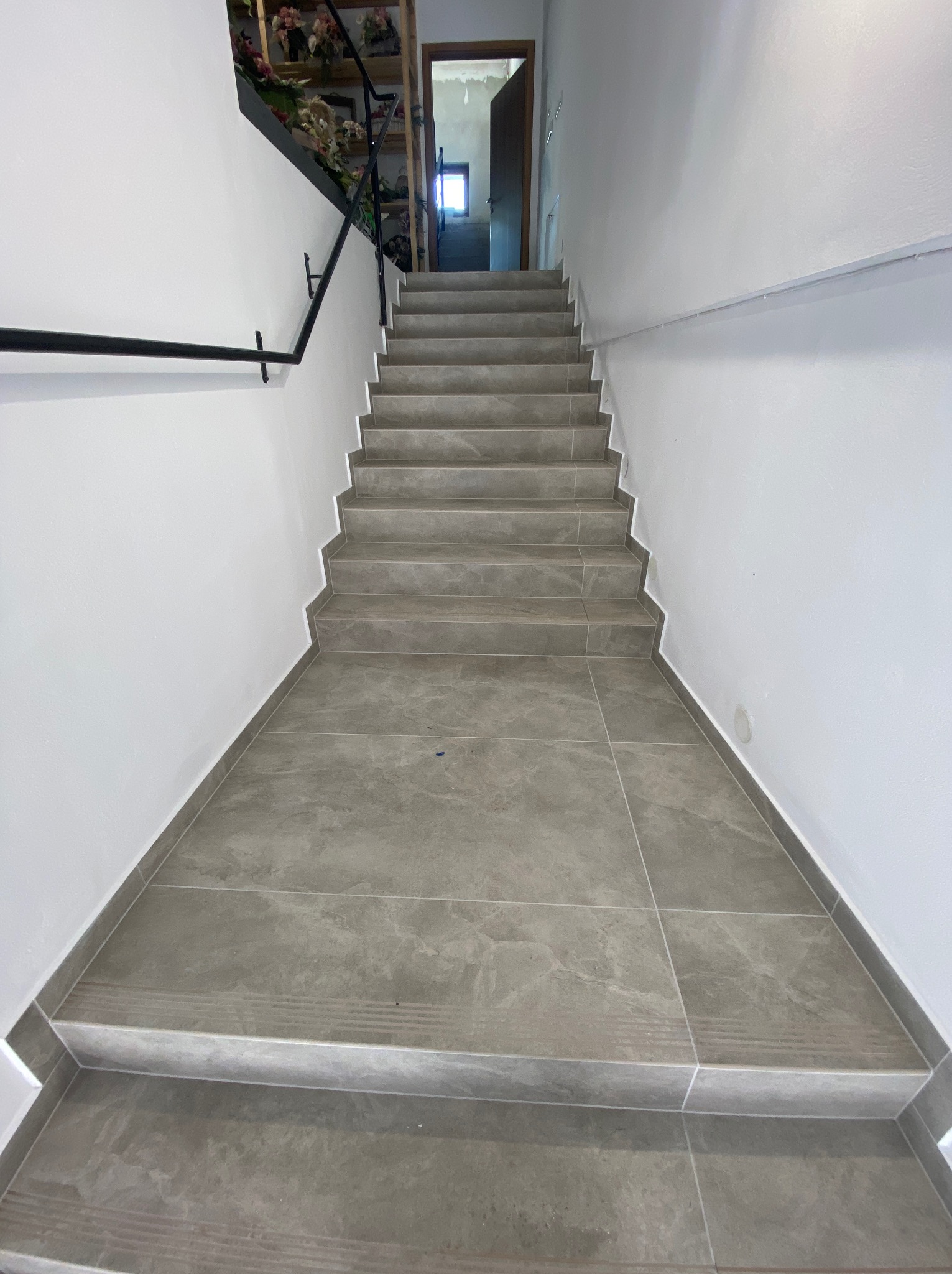 Schody wyłożone szarymi płytkami imitującymi marmur, prowadzące w górę, z czarną metalową poręczą po lewej stronie i białymi ścianami po obu stronach.