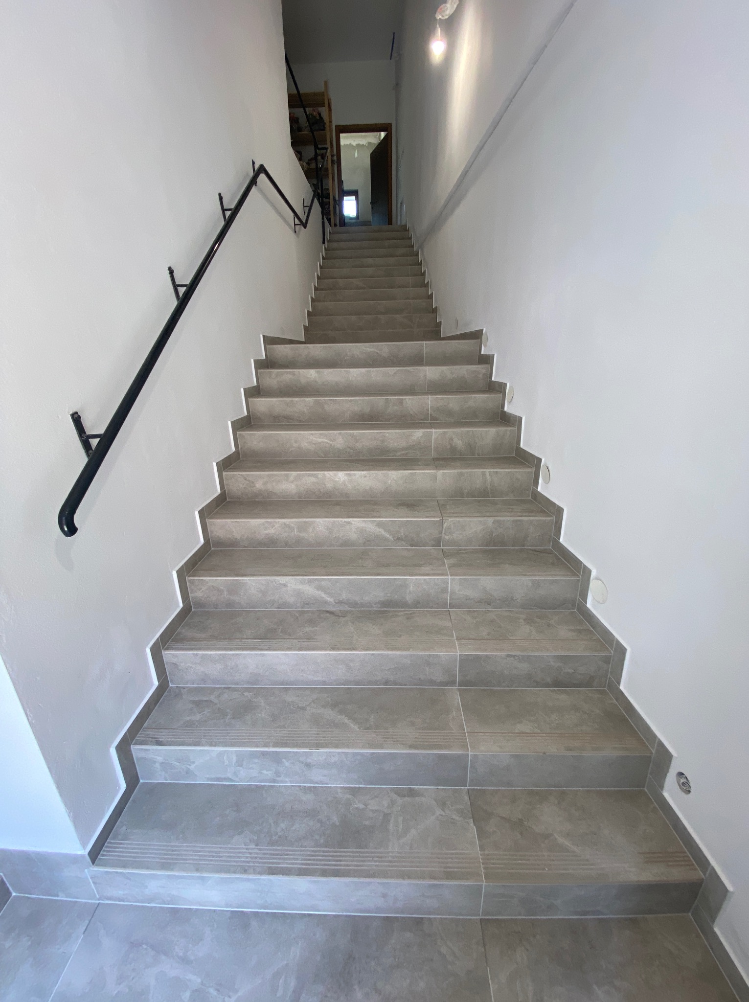 Wykończone schody wewnętrzne z szarych płytek imitujących marmur, z czarną metalową poręczą mocowaną do ściany, oświetlone punktowymi lampami LED przy podłodze.