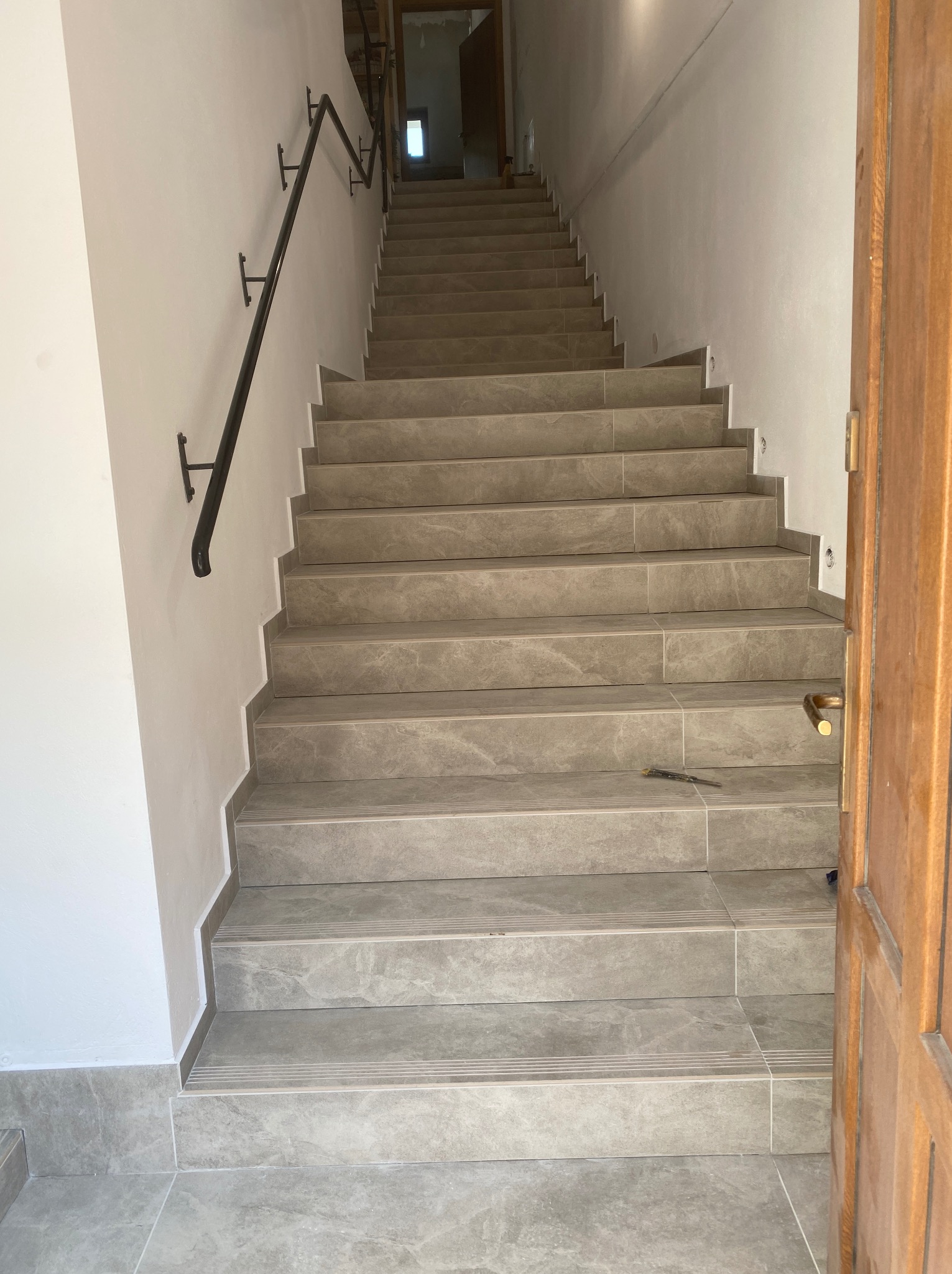 Wykończone schody wewnętrzne z szarymi płytkami imitującymi kamień, czarna metalowa poręcz na białej ścianie, widoczne narzędzie glazurnicze na jednym ze stopni, otwarte drewniane drzwi po prawej...
