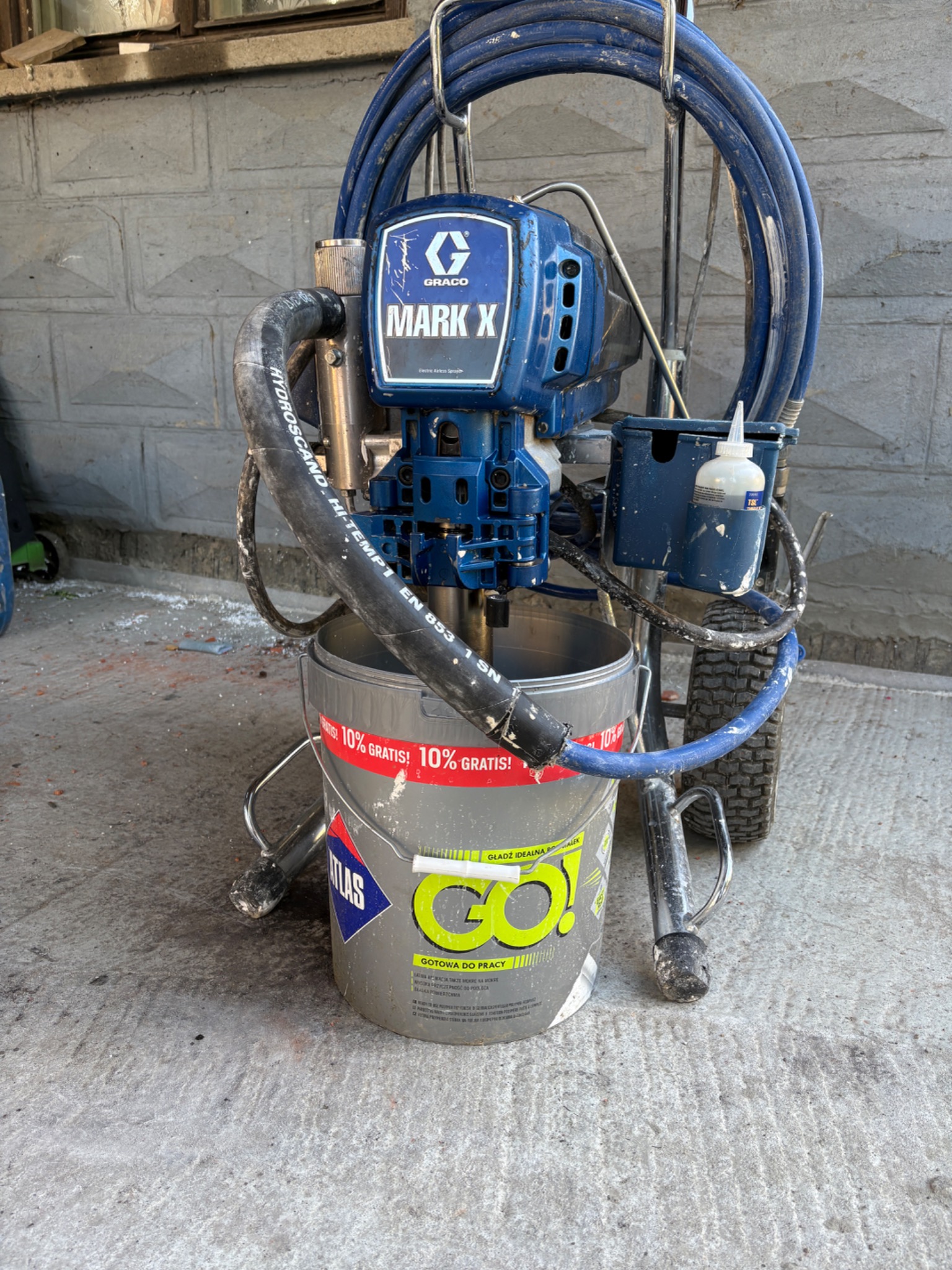 Agregat malarski Graco Mark X na wiadrze z gładzią Atlas Go! na betonowej posadzce. Widoczne przewody i akcesoria. Sprzęt gotowy do użycia.