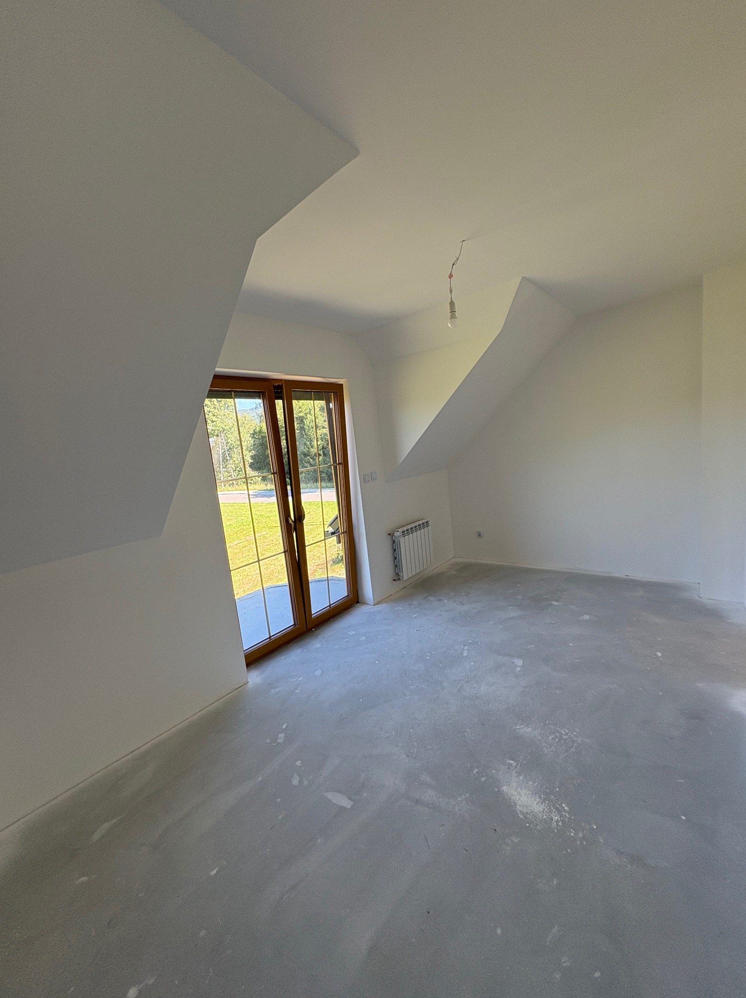 Białe poddasze po gładziowaniu, z oknem balkonowym, grzejnikiem i surową żarówką. Widoczne ślady po szlifowaniu gładzi na podłodze.