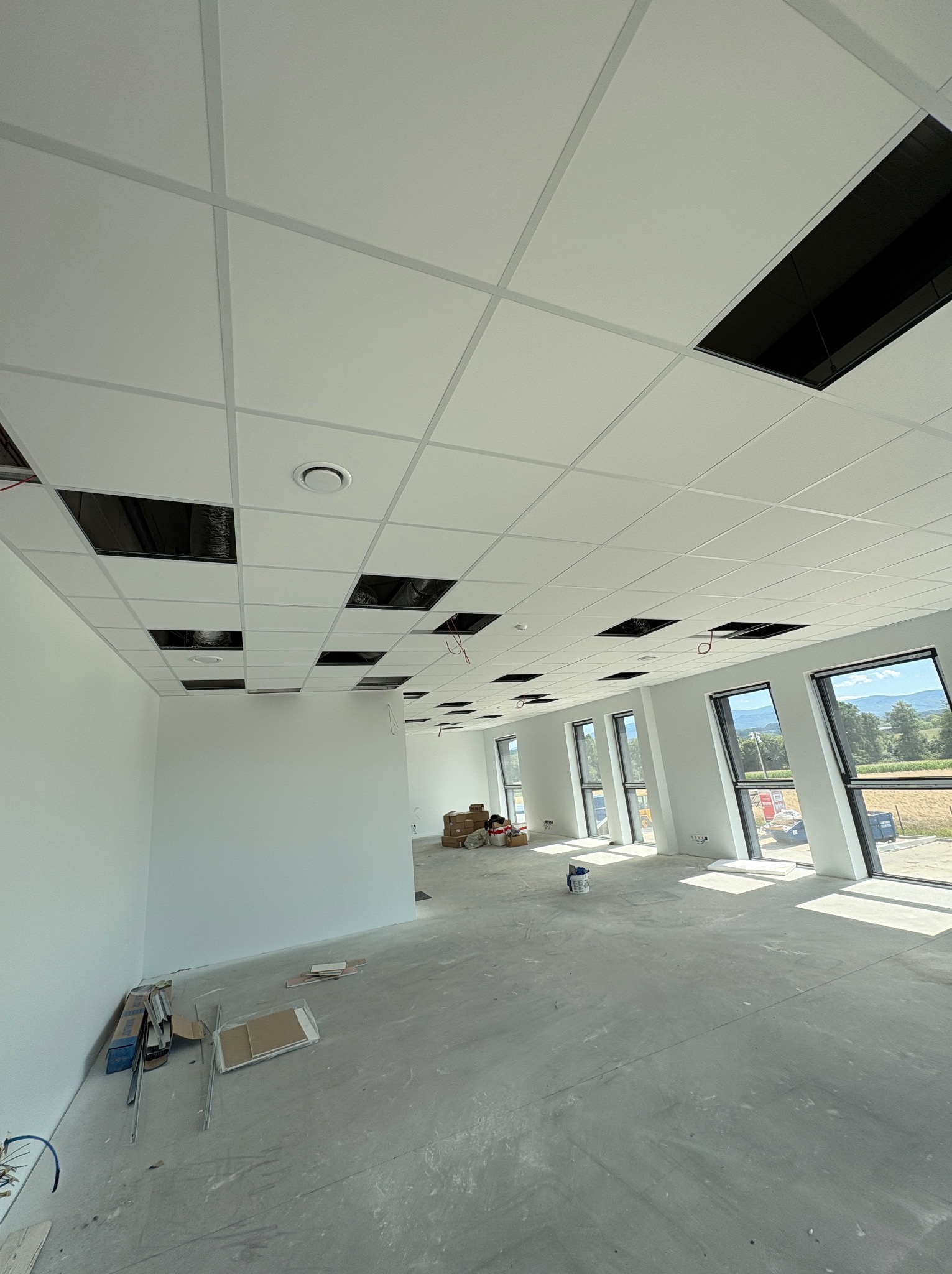 Nowoczesne wnętrze biurowe z podwieszanym sufitem kasetonowym, częściowo zamontowanym, z widocznymi otworami na instalacje. Duże okna wpuszczają naturalne światło.