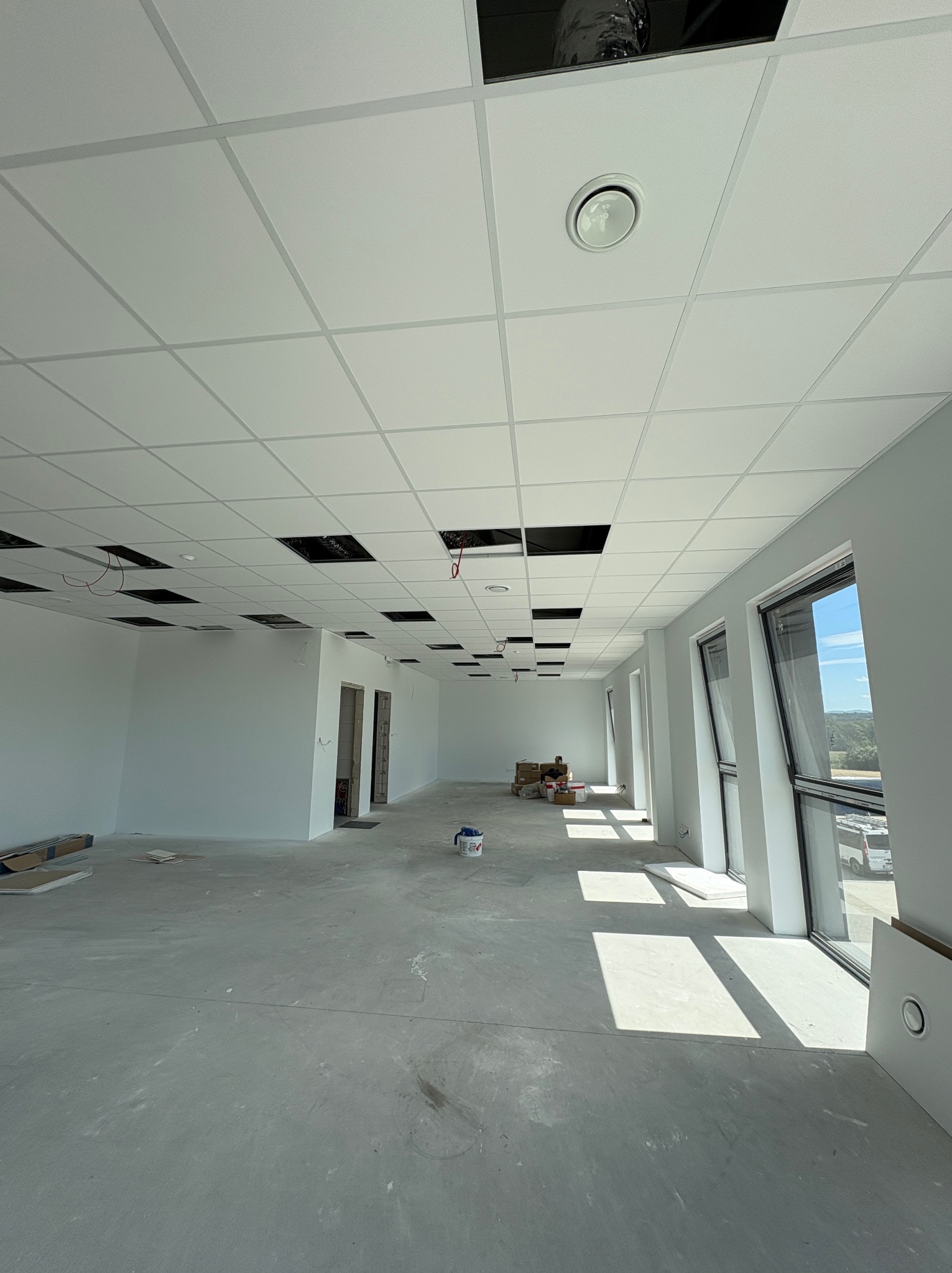 Białe, przestronne wnętrze biurowe w trakcie wykańczania: widoczne okna, podwieszany sufit z otworami, ściany po gładziowaniu i podkładzie, przygotowane do dalszych prac.