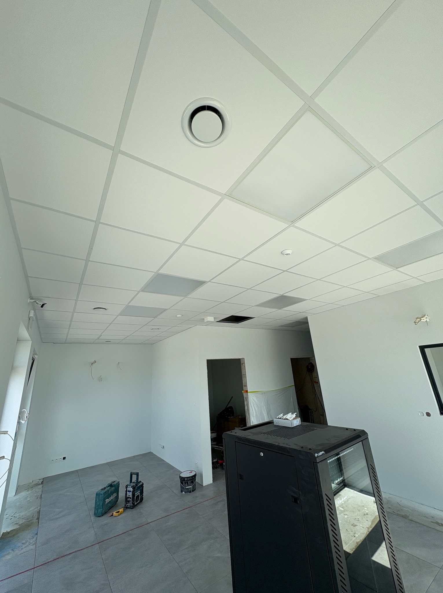 Biały sufit kasetonowy z okrągłym nawiewnikiem w nowoczesnym biurze, szare płytki podłogowe, narzędzia i szafa rack w tle, remont w trakcie.
