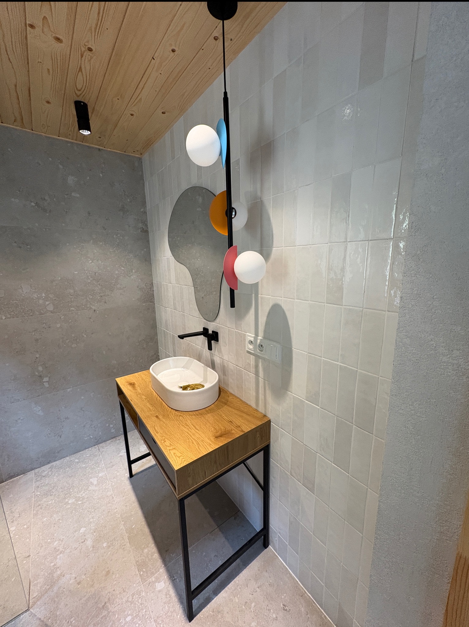 Minimalistyczna łazienka w Żywcu z umywalką na drewnianym blacie, lustrem o nieregularnym kształcie i białymi płytkami na ścianie, oświetlona wiszącą lampą.