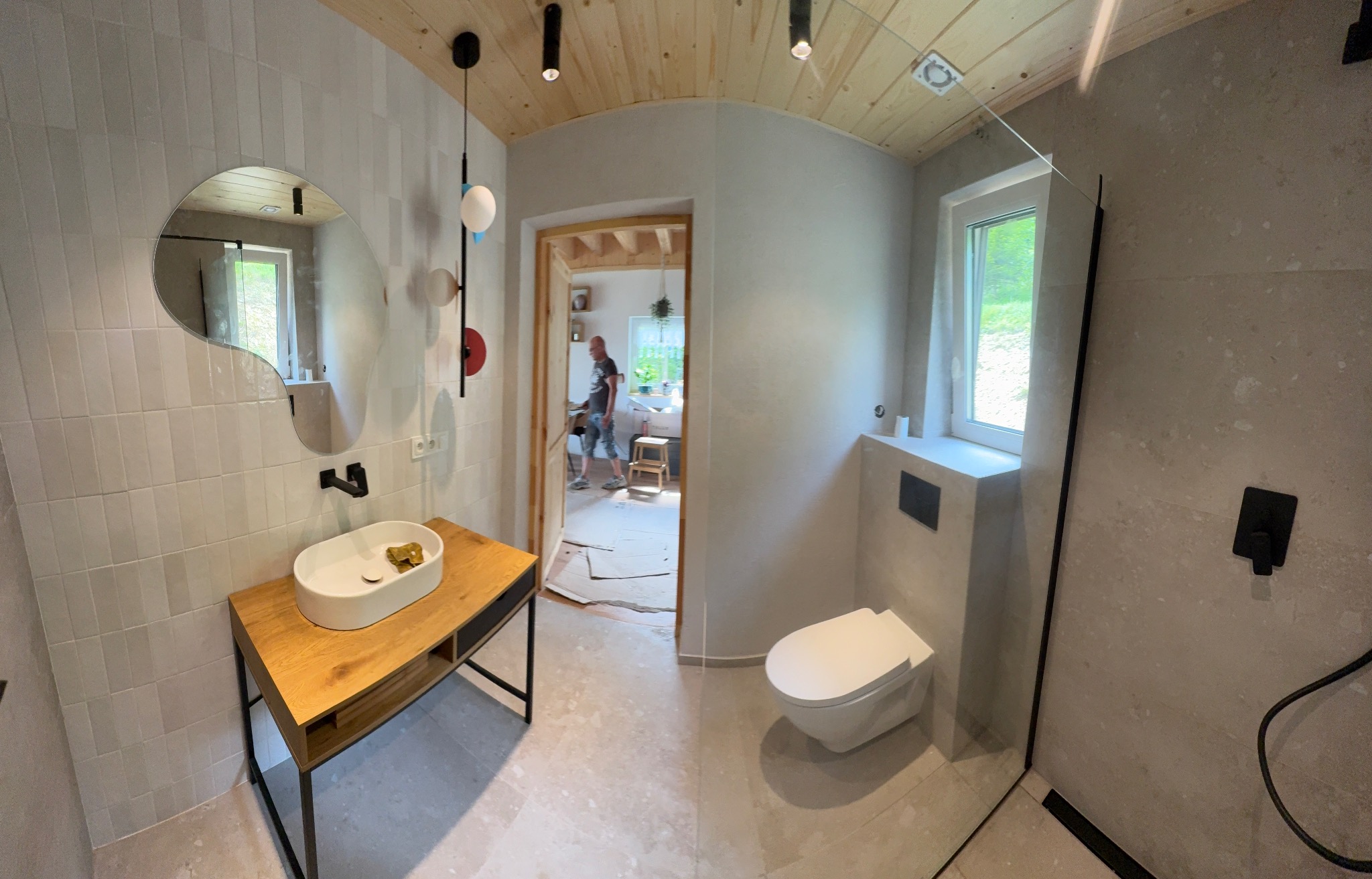 Wykończona łazienka z drewnianym sufitem, umywalką na drewnianym blacie i kabiną prysznicową. W tle widoczny fragment innego pomieszczenia w trakcie remontu.