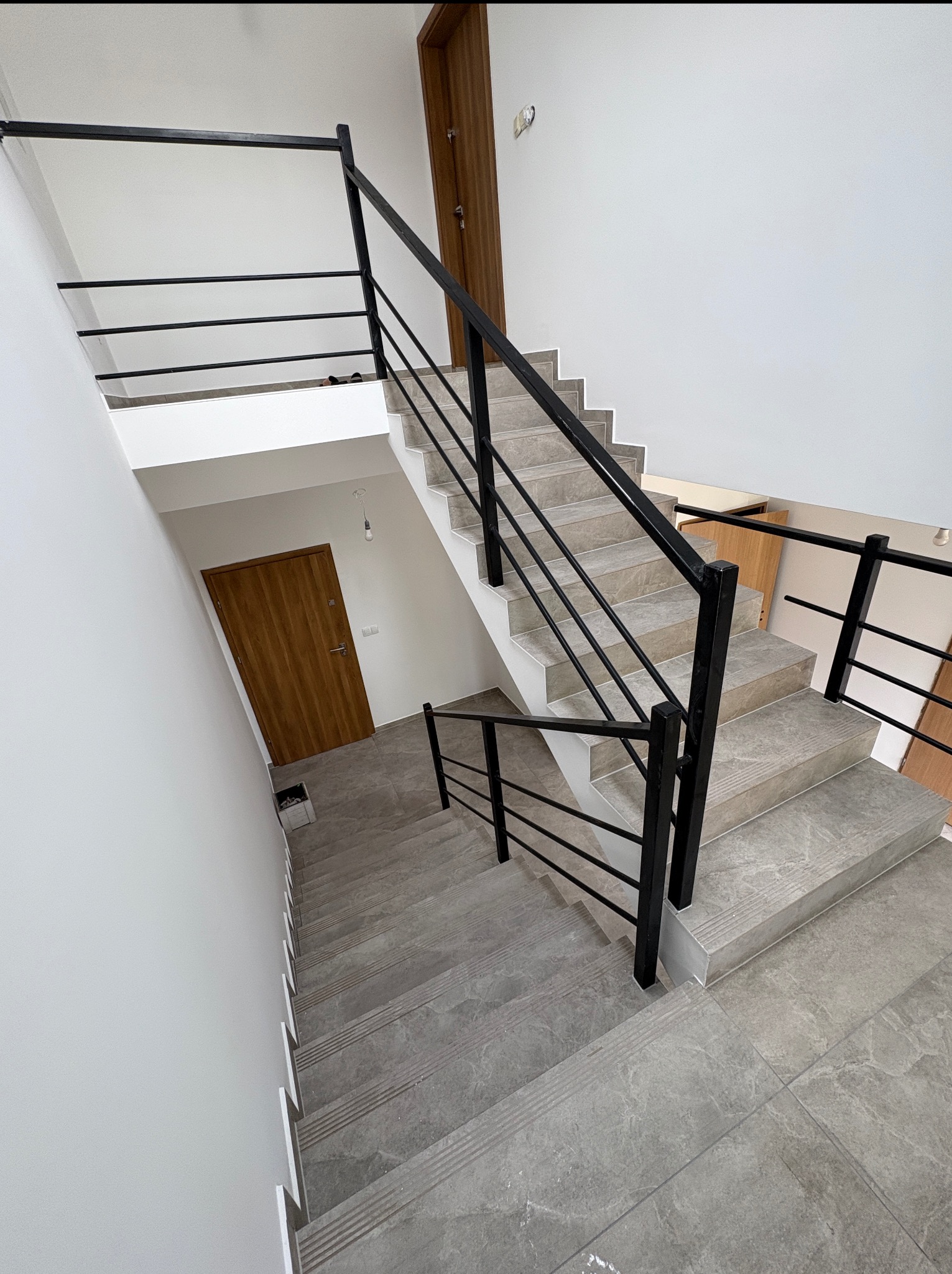 Klatka schodowa z szarymi płytkami imitującymi marmur i czarną, metalową balustradą. Widok z góry na dół, dwie drewniane drzwi w tle.