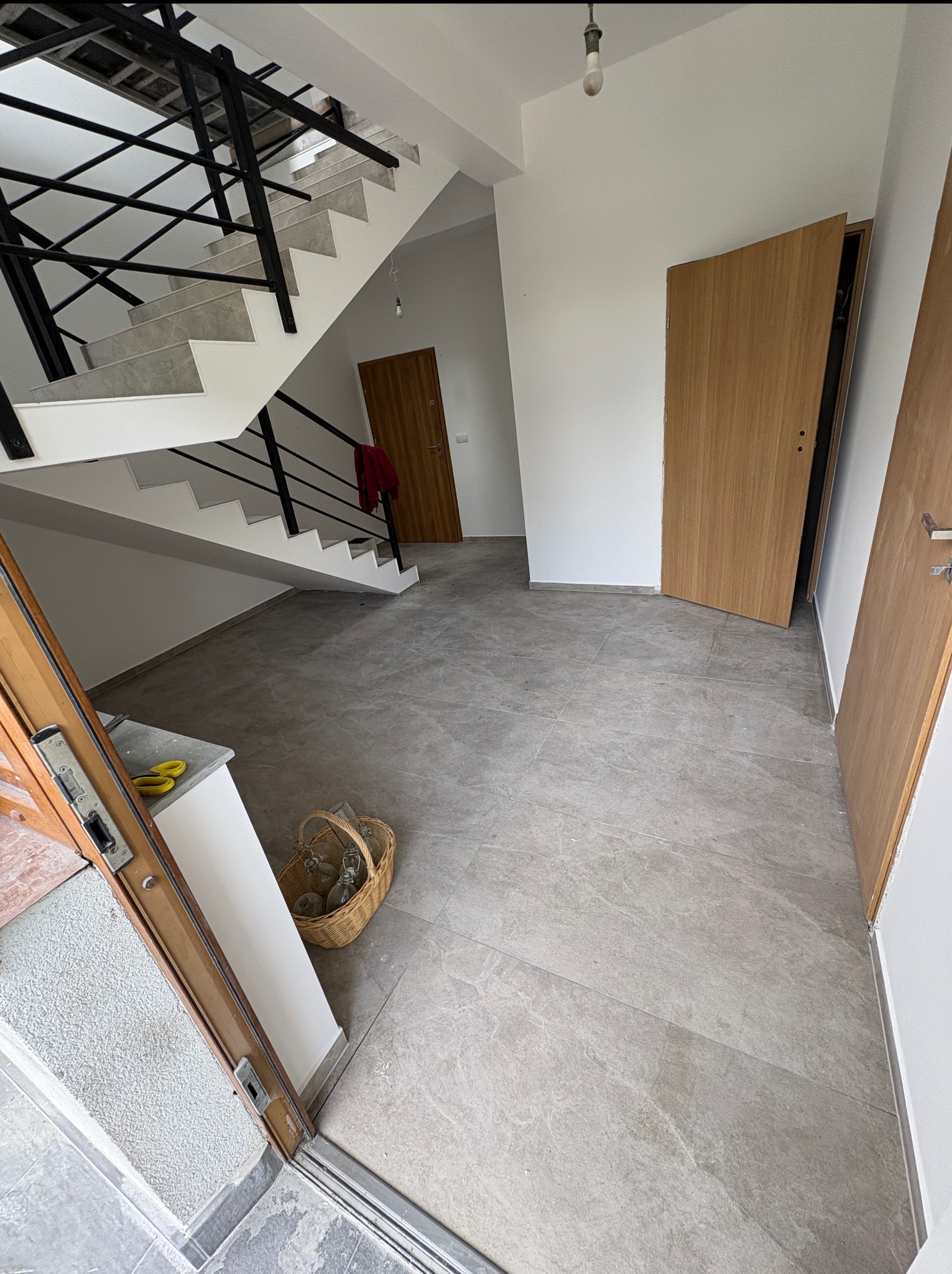 Wykończone wnętrze z szarymi płytkami podłogowymi, białymi ścianami, schodami z czarną balustradą i drewnianymi drzwiami. Koszyk z butelkami przy wejściu.