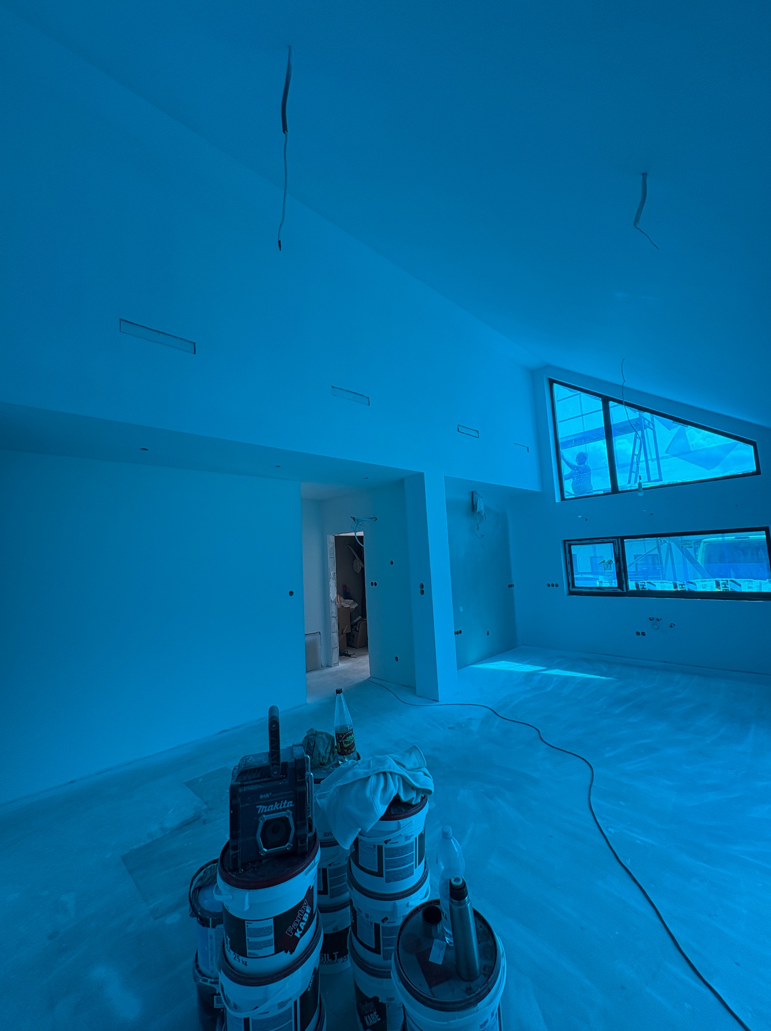 Wnętrze po malowaniu: białe ściany, okna z widokiem na pracownika na rusztowaniu. Na pierwszym planie wiaderka z farbą i narzędzia malarskie.