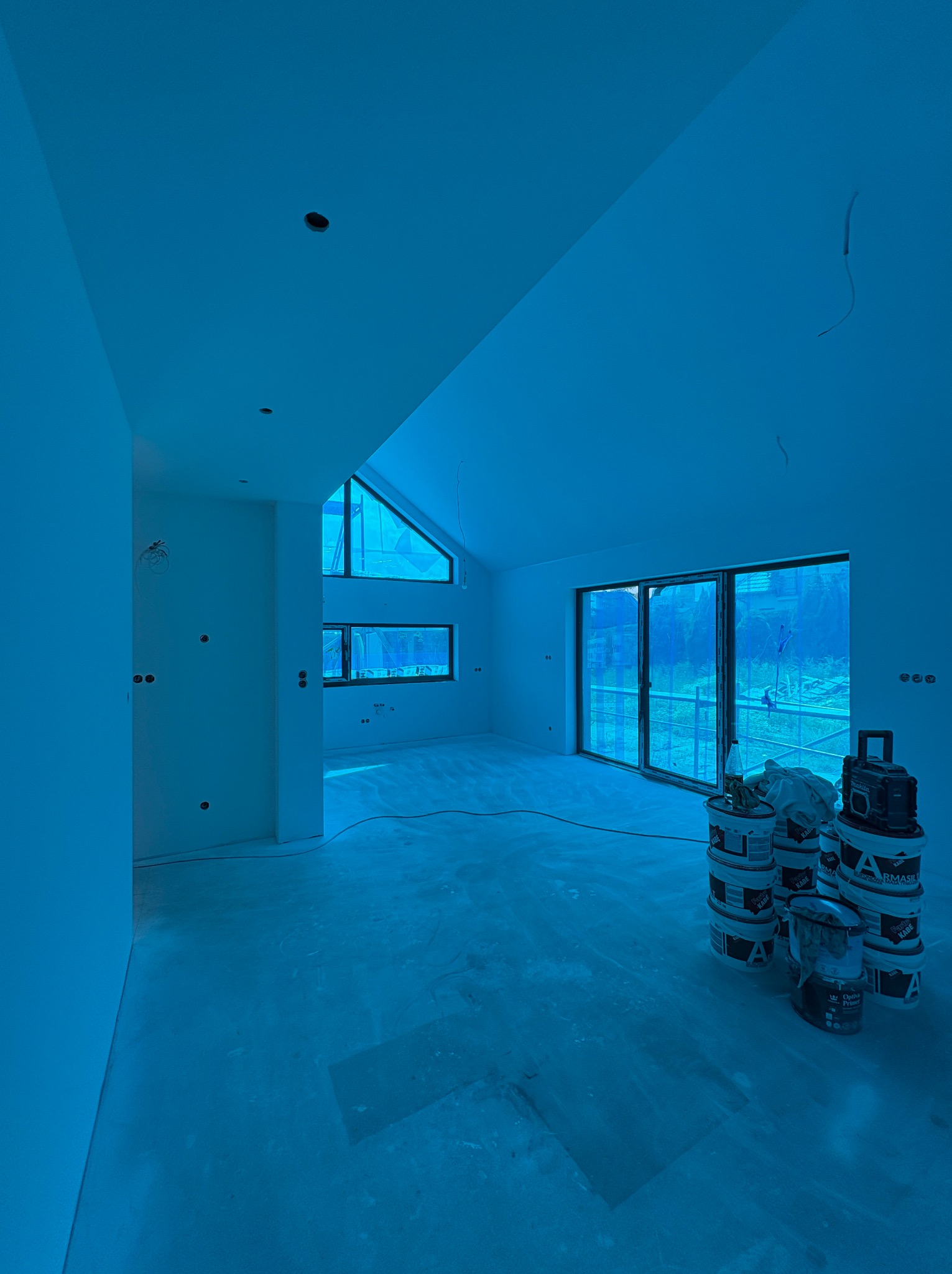 Wnętrze po gładziowaniu, przygotowane do malowania. Widoczne ściany, okna, puszki elektryczne i wiaderka z farbą. Skosy sufitu i duże okno dachowe doświetlają pomieszczenie.