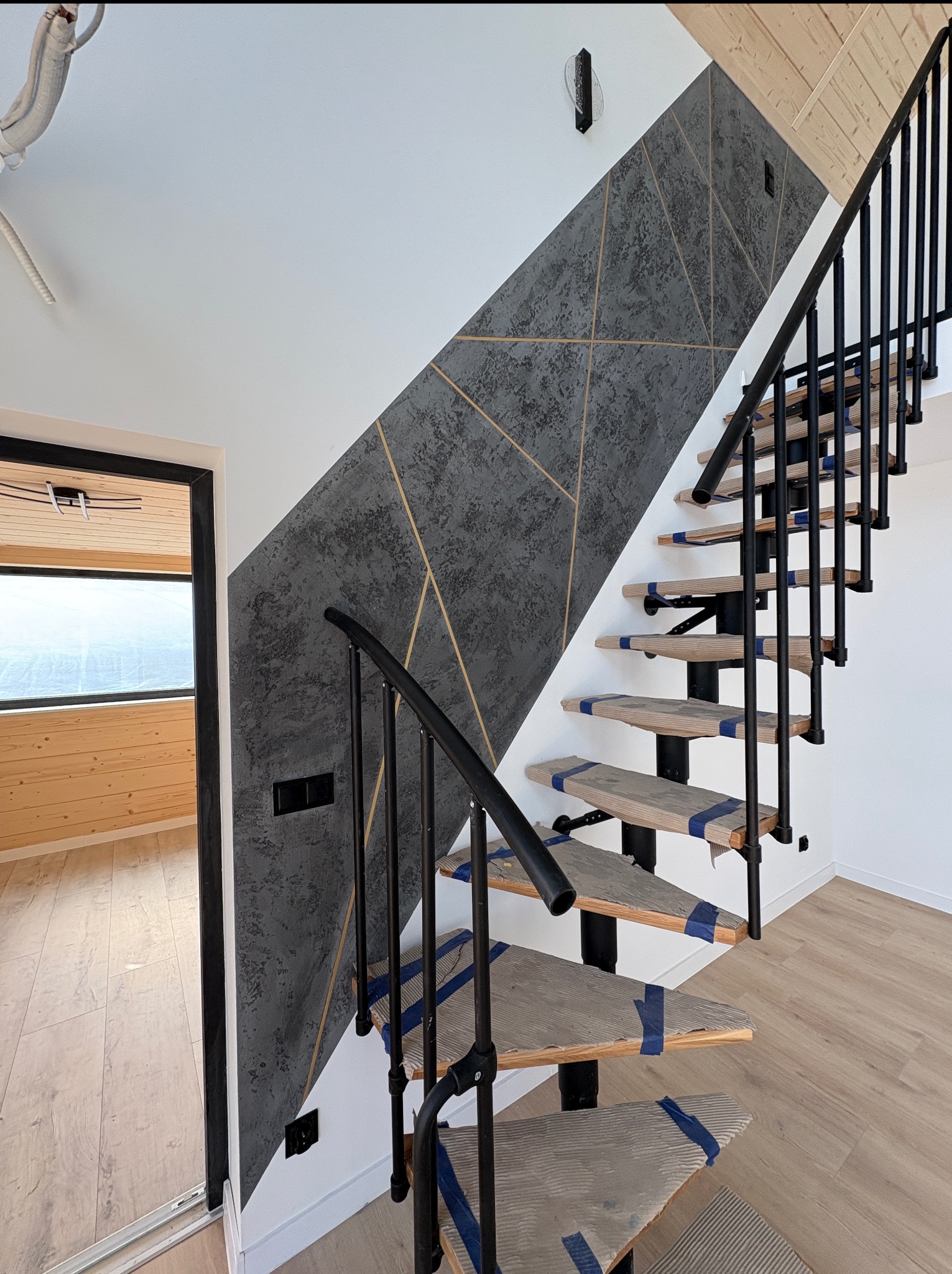 Ściana z dekoracyjną szarą strukturą i złotymi liniami, schody z czarną balustradą zabezpieczone taśmą malarską, widok na drewniane wnętrze przez otwarte drzwi.