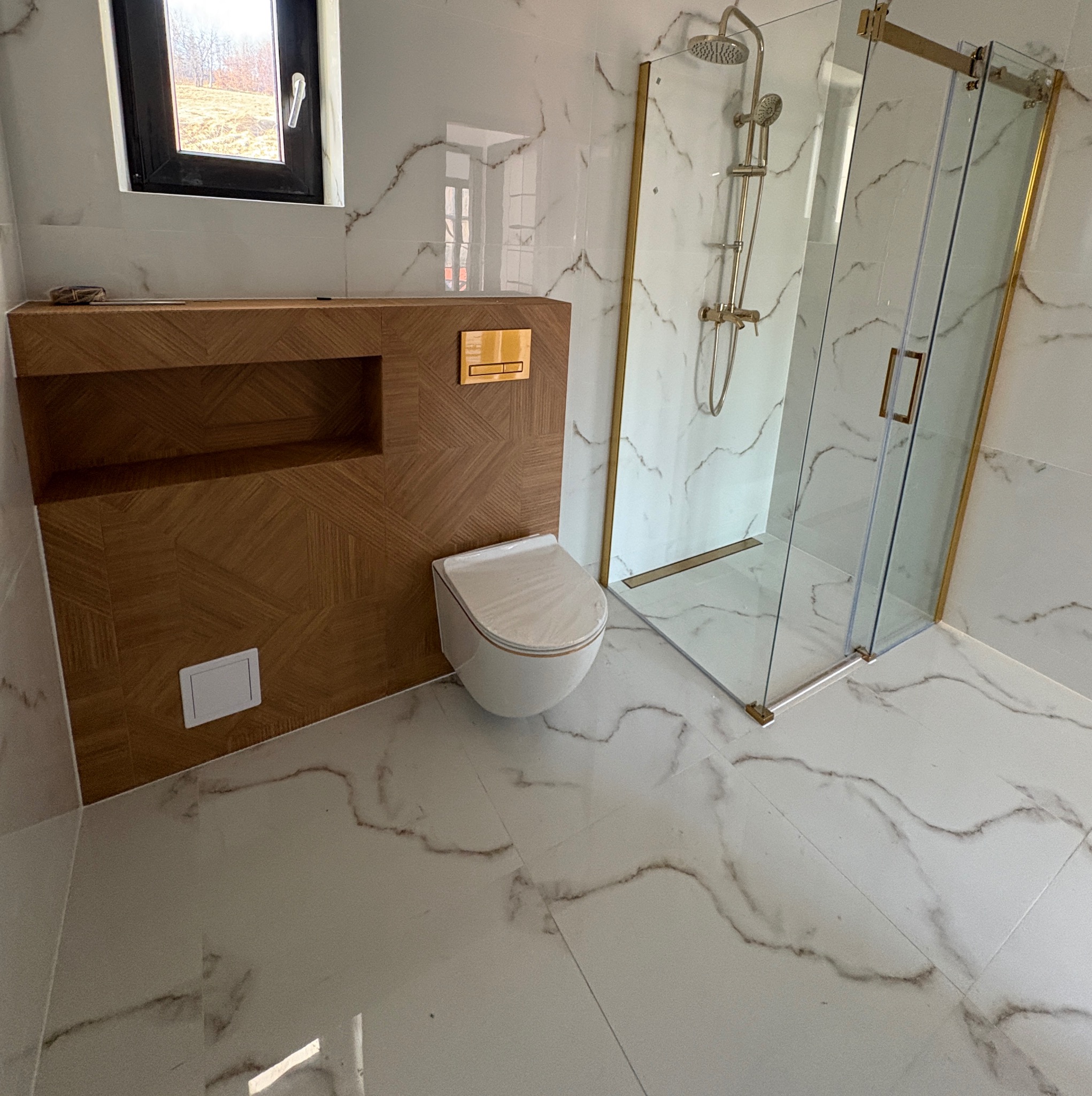 Nowoczesna łazienka z biało-marmurowymi płytkami, kabiną prysznicową ze złotymi detalami i zabudową WC z drewnianym panelem dekoracyjnym.