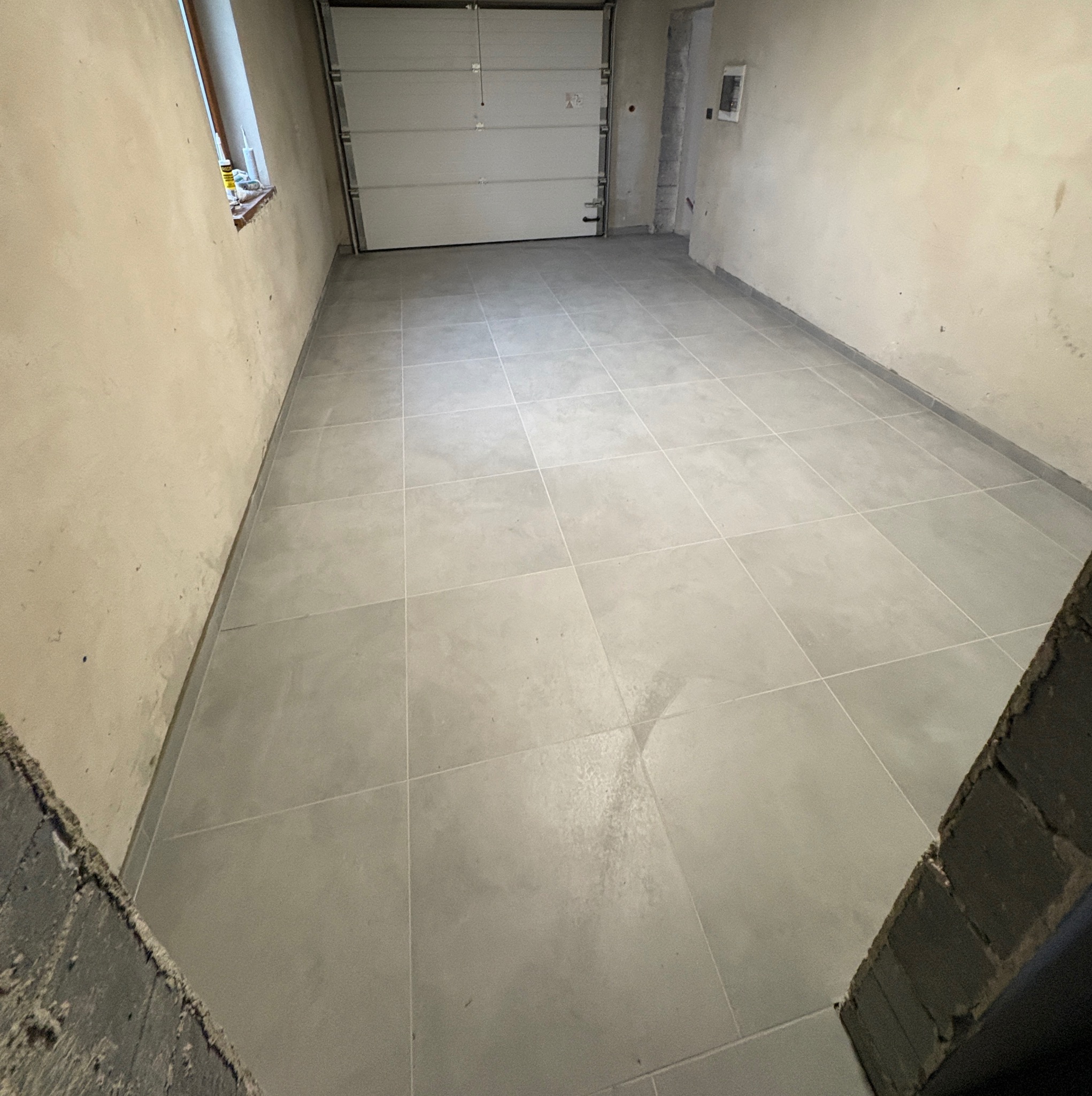 Wykończone wnętrze garażu z jasnoszarą, kwadratową podłogą z płytek. Ściany surowe, w tle brama garażowa i okno. Widoczne narzędzia na parapecie.