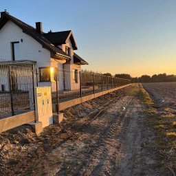 LuxPłot Piotr Szczygielski - Długa linia nowoczesnego, czarnego ogrodzenia z siatki, na betonowej podmurówce, otacza dom z numerem 13c, widok w kierunku pola uprawnego oświetlonego zachodzącym słońcem.