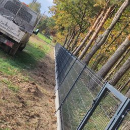 LuxPłot Piotr Szczygielski - Nowo zamontowane szare ogrodzenie panelowe z furtką, biegnące wzdłuż granicy działki z młodym lasem, obok zaparkowany samochód dostawczy firmy geodezyjnej.