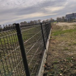 LuxPłot Piotr Szczygielski - Długi fragment czarnego ogrodzenia z siatki panelowej, zamontowany na betonowej podmurówce, ciągnący się wzdłuż nierównego, lekko trawiastego terenu z widocznymi liśćmi, z domem w tle, w pochmurny...