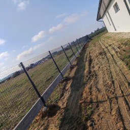 LuxPłot Piotr Szczygielski - Długa linia ogrodzenia z siatki, osadzona na betonowej podmurówce, ciągnąca się wzdłuż nierównego, piaszczystego terenu obok nowego budynku w słoneczny dzień.