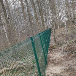 LuxPłot Piotr Szczygielski - Długi odcinek zielonego ogrodzenia z siatki, biegnący wzdłuż zalesionego zbocza, z widocznymi słupkami i ziemią po drugiej stronie.