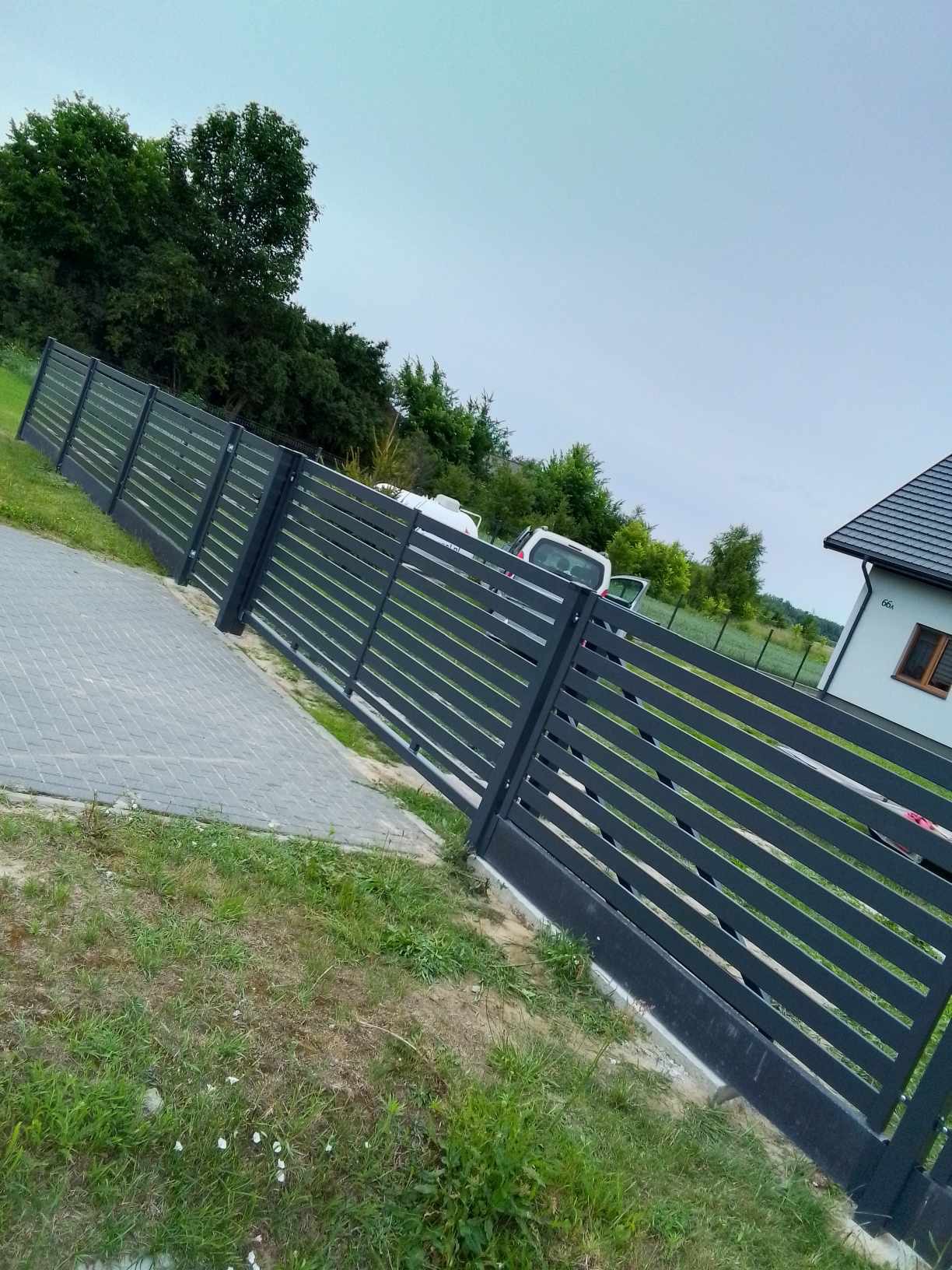 Nowoczesne, poziome przęsła grafitowej konstrukcji ogrodzeniowej, widoczne z perspektywy chodnika z szarej kostki, z domem i samochodem w tle. Uwagę zwraca świeżo wylany beton u podstawy słupków.
