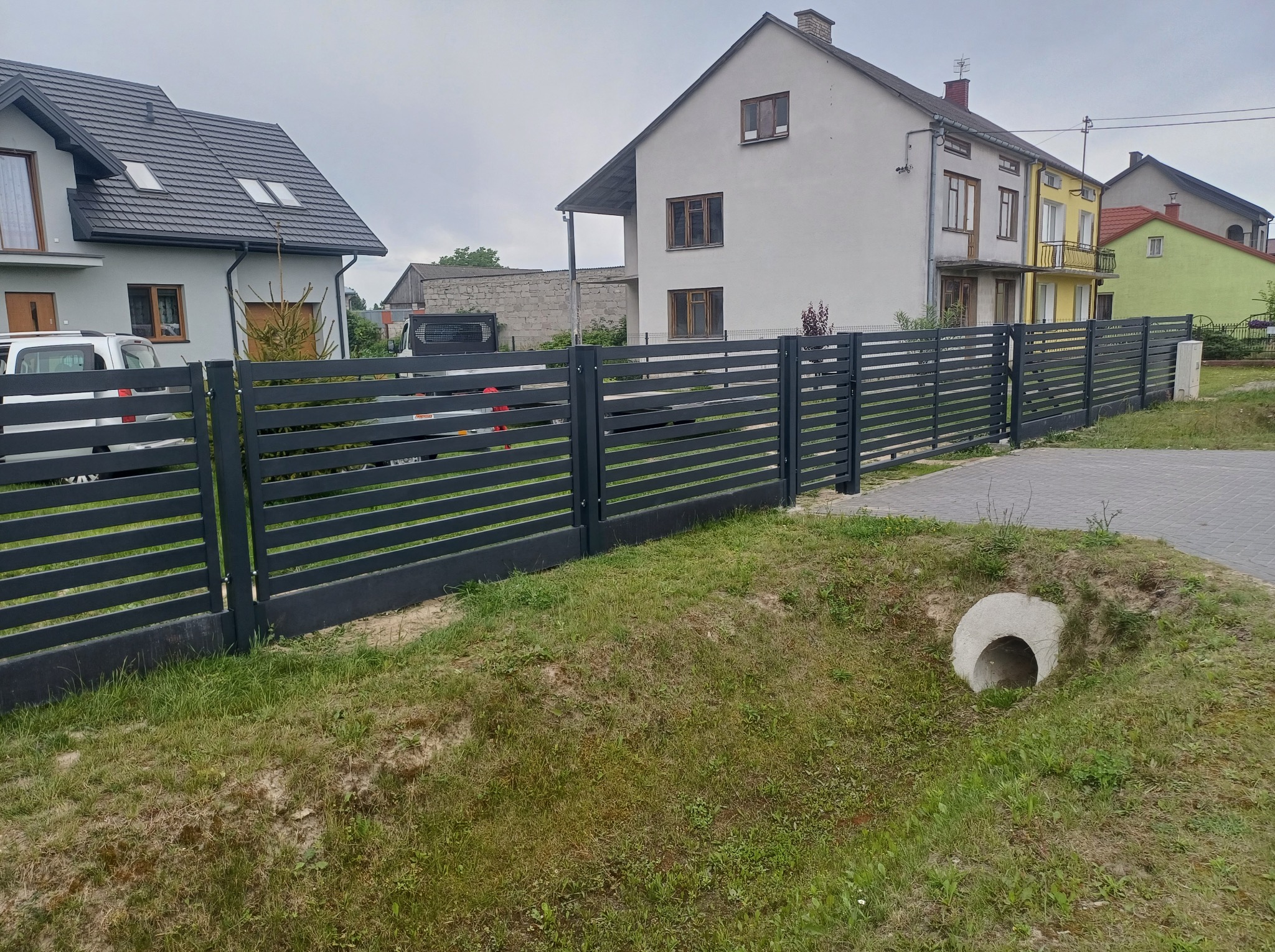Nowoczesne, poziome, szare ogrodzenie z metalowych profili, oddzielające posesje z domami o zróżnicowanej architekturze i kolorystyce, widoczne ujście betonowej rury drenażowej.