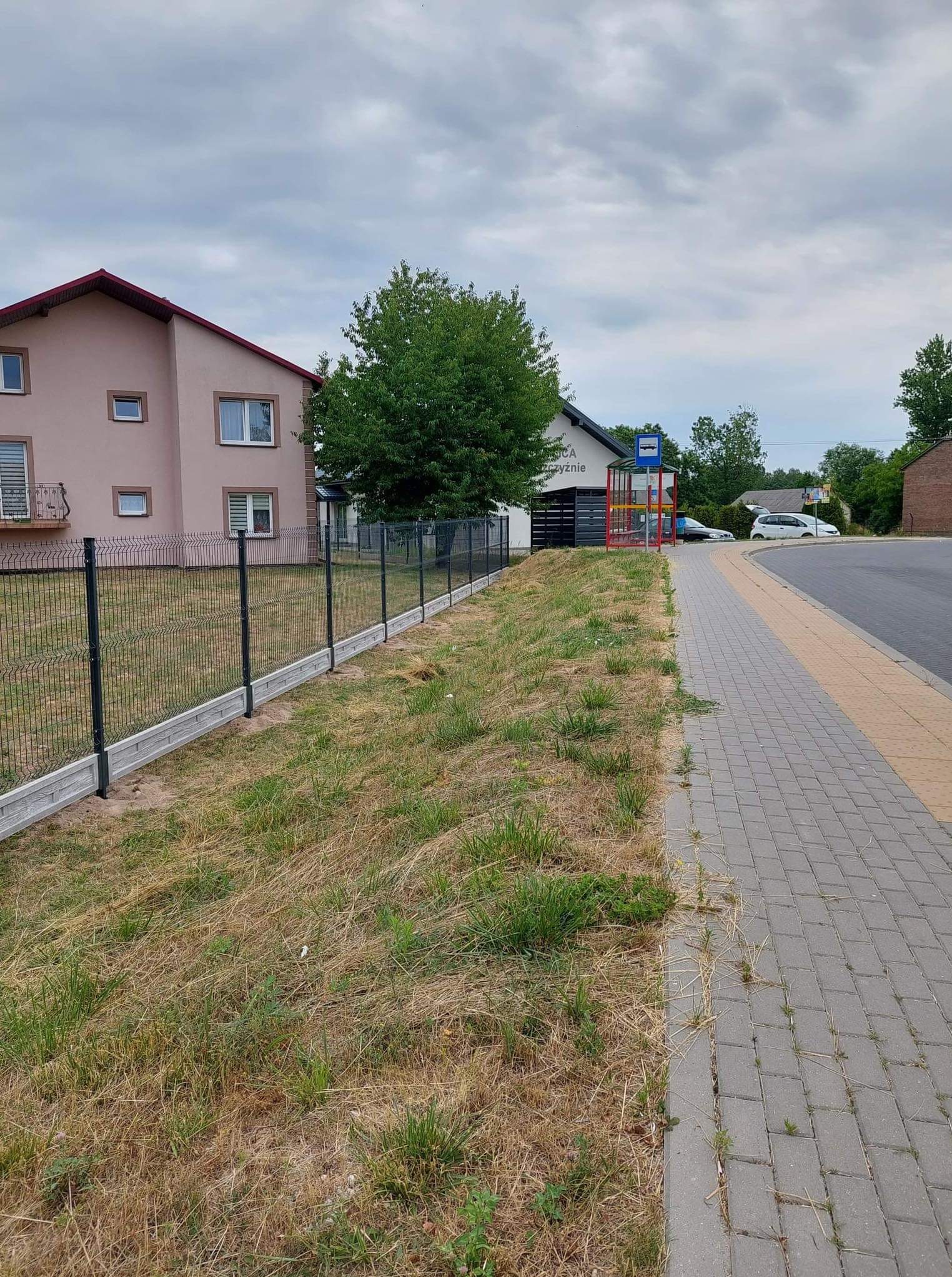 Czarny panelowy system ogrodzeniowy z betonową podmurówką, oddzielający posesję od chodnika i przystanku autobusowego w pochmurny dzień.