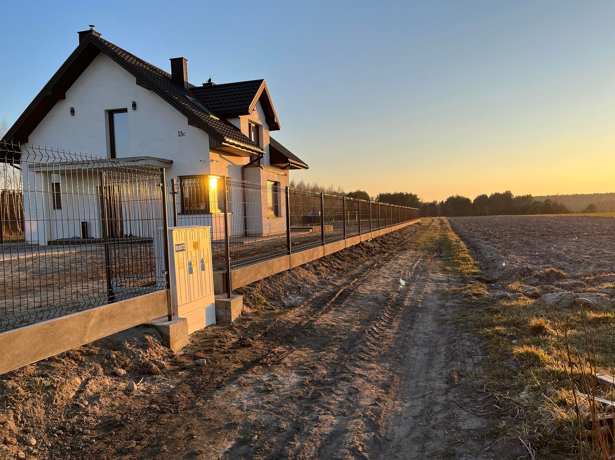 Długa linia nowoczesnego, czarnego ogrodzenia z siatki, na betonowej podmurówce, otacza dom z numerem 13c, widok w kierunku pola uprawnego oświetlonego zachodzącym słońcem.