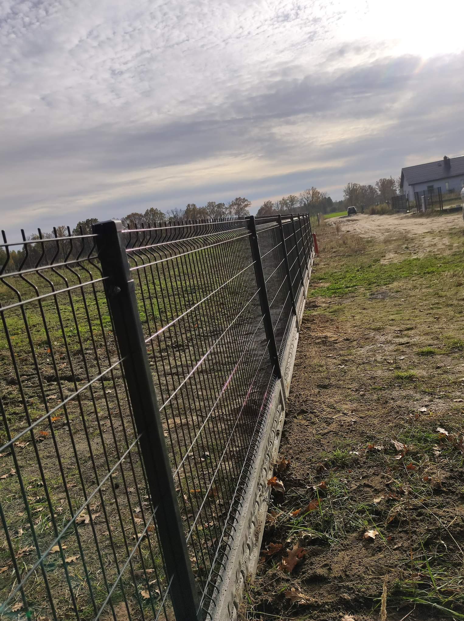 Długi fragment czarnego ogrodzenia z siatki panelowej, zamontowany na betonowej podmurówce, ciągnący się wzdłuż nierównego, lekko trawiastego terenu z widocznymi liśćmi, z domem w tle, w pochmurny...