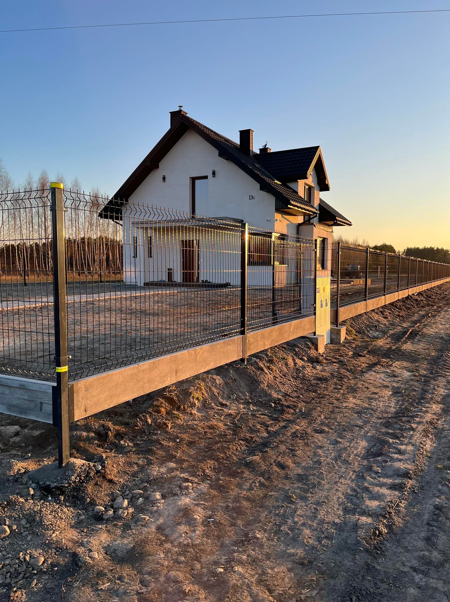 Nowoczesne ogrodzenie panelowe z czarnych paneli na betonowej podmurówce, otaczające dom jednorodzinny w słoneczny poranek.