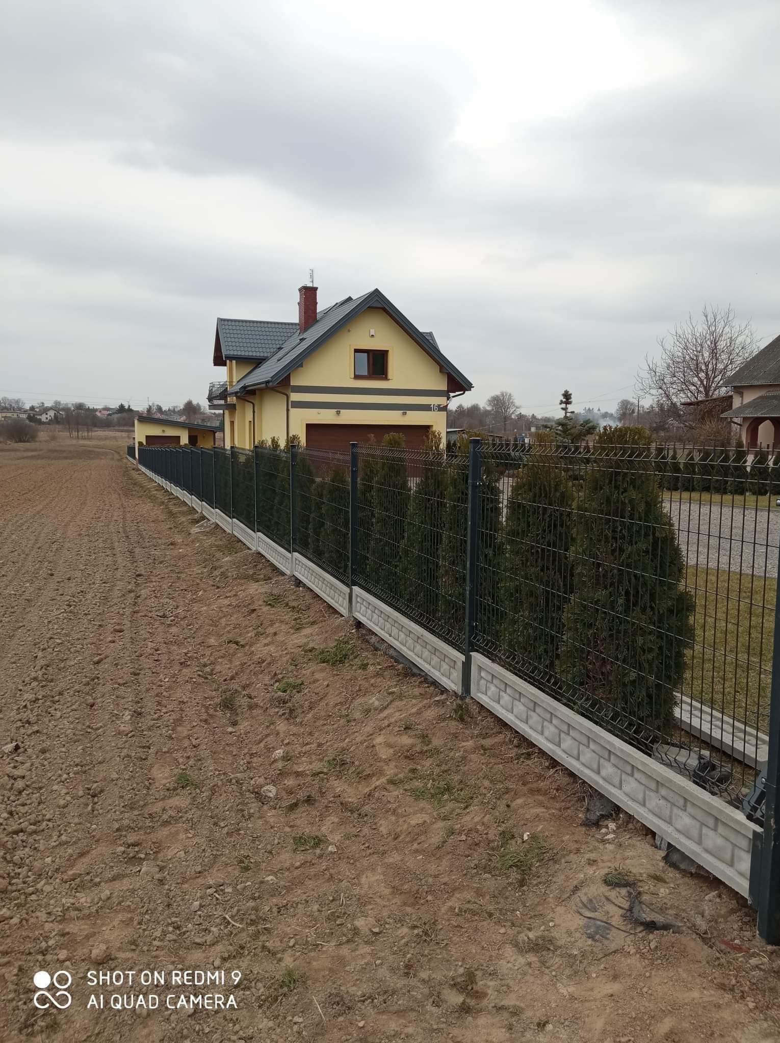 Długa, ciemna siatka ogrodzeniowa z podmurówką imitującą cegłę, oddzielająca pole od posesji z żółtym domem i tujami, pochmurne niebo.