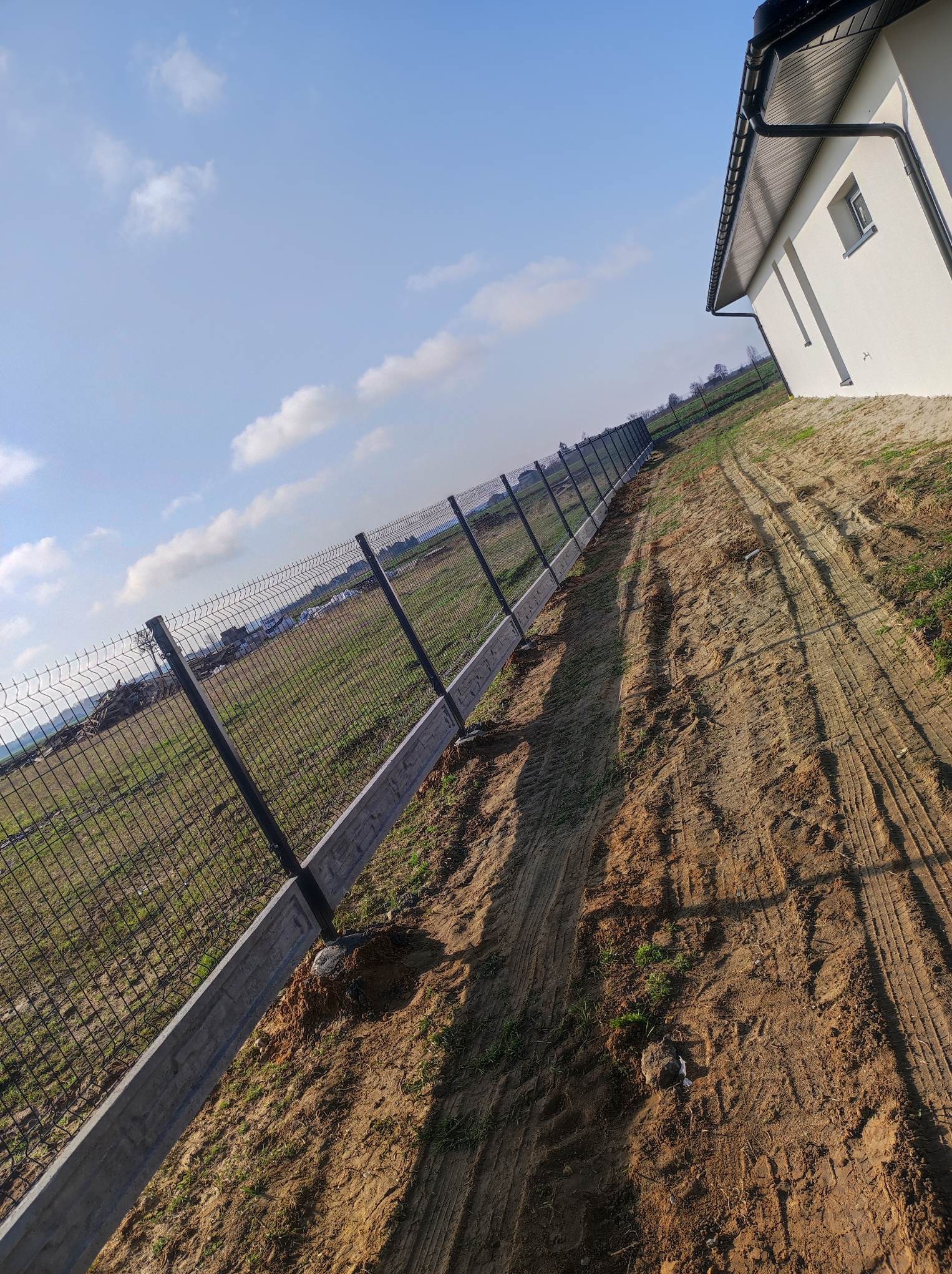 Długa linia ogrodzenia z siatki, osadzona na betonowej podmurówce, ciągnąca się wzdłuż nierównego, piaszczystego terenu obok nowego budynku w słoneczny dzień.