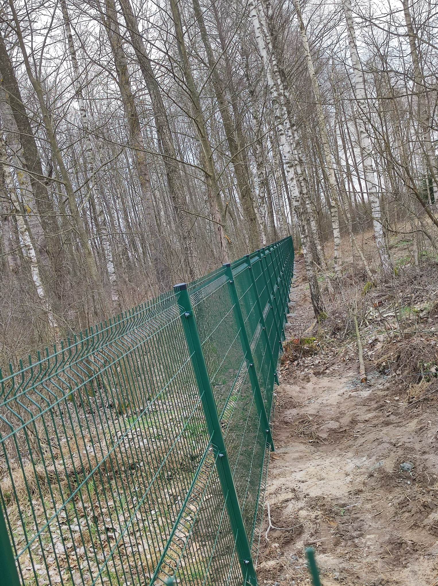 Długi odcinek zielonego ogrodzenia z siatki, biegnący wzdłuż zalesionego zbocza, z widocznymi słupkami i ziemią po drugiej stronie.