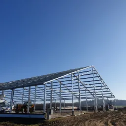 Stalowy szkielet hali w trakcie budowy, widoczny na tle błękitnego nieba i lasu, z zaparkowanym podnośnikiem koszowym i traktorem na placu budowy.