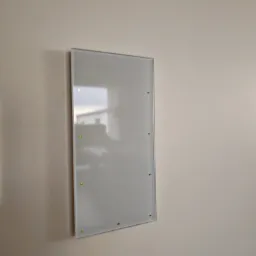 Szklany panel dotykowy, prawdopodobnie sterownik systemu inteligentnego domu, zamontowany na kremowej ścianie, z widocznymi zielonymi diodami LED.