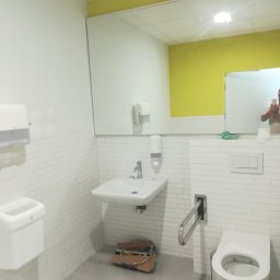 DAST - Biała łazienka w trakcie montażu armatury, widoczna umywalka, toaleta z uchwytem dla niepełnosprawnych, dozowniki i lustro z odbiciem osoby robiącej zdjęcie.