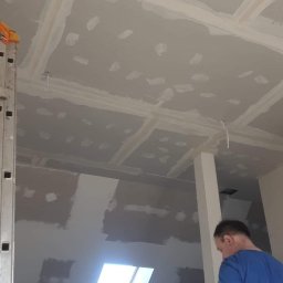 DAST - Poddasze w trakcie prac, widoczne łączenia płyt gipsowo-kartonowych wypełnione masą szpachlową, pracownik stoi na drabinie, w tle okno dachowe.