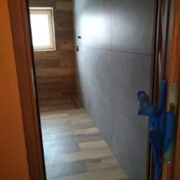 Nowoczesna łazienka z drewnopodobnymi płytkami na podłodze i ścianie, szare płytki na ścianie naprzeciwko, widok zza częściowo otwartych drewnianych drzwi, niebieska szmatka na klamce.