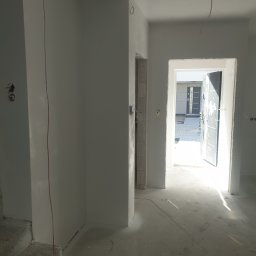 DAST - Wnętrze w trakcie prac wykończeniowych: świeżo położone gładzie na ścianach, widoczne kable elektryczne, betonowe schody i surowa podłoga z resztkami taśmy malarskiej, otwarte drzwi wejściowe w tle.